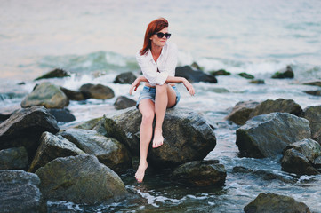 Молодая красивая девушка в очках сидит на камнях на берегу моря 