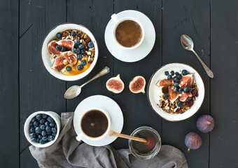 Obraz na płótnie Canvas Healthy breakfast set. Bowls of oat granola with yogurt, fresh blueberries and figs, coffee, honey, over black wooden backdrop