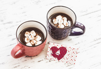 Two cups of hot chocolate with marshmallows and  Valentine's day poscard on bright wooden surface