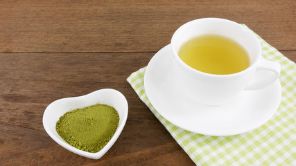 The Japanese matcha green tea powder on ceramic heart shaped bowl and cup of hot green tea with cotton fabric on wooden planks.