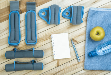 Sport Equipment. Dumbbells,  Ankle Weights, Wrist Weights, Towel, Apple, Bottle Of Water And Notebook To Workout Plan On Boards. Sport Fitness Background