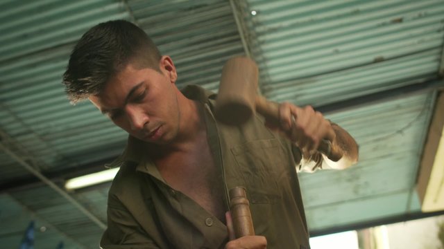 Young man at work as sculptor learning craftsman profession, working with hammer and chisel. The artist carves a raw block of wood to make a wooden statue