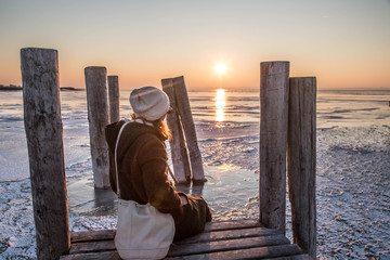 Frau sitzt am Holzsteg und blickt auf gefrorenen See bei Sonnenuntergang