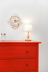 Room interior with red wooden commode, lamp and clock on light wall background