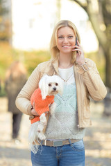Happy young woman talking on the phone and carrying her cute little dog while walking in a park
