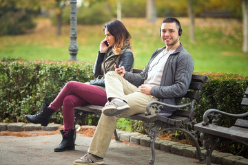 Young man listening to music while woman sitting on the same park bench is talking on the phone