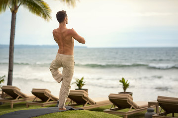 young man making yoga exercises on beach from back