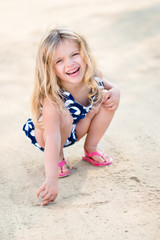 Beautiful laughing little girl with long blond hair squatting and drawing in the sand on the beach...