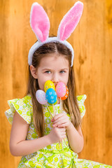 Portrait of smiling little girl with long blond hair wearing pink and white rabbit or bunny ears and holding bunch of painted colorful eggs. Easter celebrations. Wooden background. Studio portrait