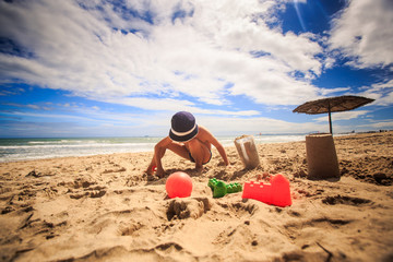 Little Boy in Hat Fills Plastic Mug with Sand Make Cake on Beach