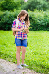 Pretty teenager girl in casual clothes with backpack holding digital tablet in her hand, typing and reading, outdoor portrait