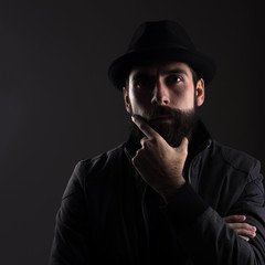 High contrast portrait of serious bearded man wearing black hat thinking looking at camera over black background.