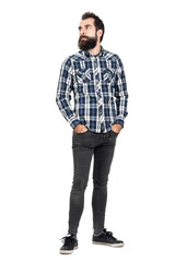 Fototapeta premium Serious bearded hipster in checked plaid shirt with hands in pockets looking away. Full body length portrait isolated over white studio background.