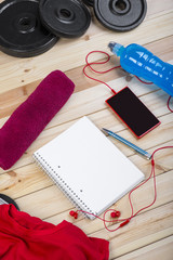Sport Equipment. Weights, Red Shirt, Isotonic Drink, Towel, Smart Phone With Earphones And Notebook To Workout Plan On Wooden Table. Sport Fitness Background