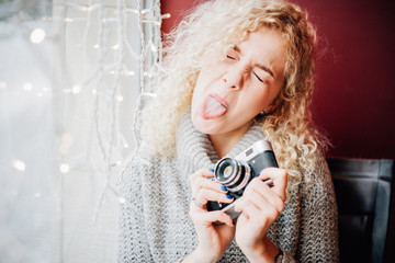 Young blond curly female in warm sweater holding old film camera, ape and smiling in the cafe, winter city outside the window