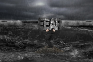 Businessman carrying old structure of FEAR on rock in dark ocean