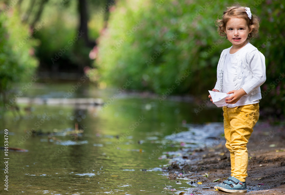 Wall mural Cute little girl runs a paper boat in the stream in the park. Stretching her hand and reaching the little ship