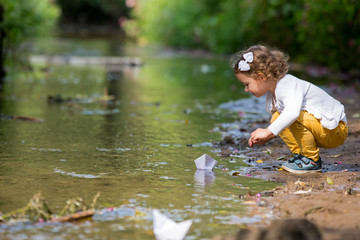 Cute little girl runs a paper boat in the stream in the park. Stretching her hand and reaching the...