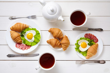 Heart shaped fried eggs, salad, croissants, salami sausage, composition and tea on white wooden table background.