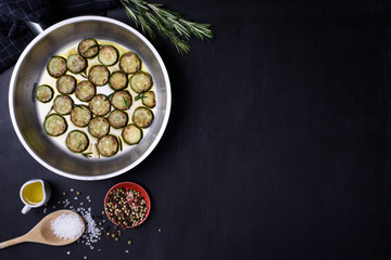 Zucchini and rosemary fried in a pan, salt and pepper over dark wooden background. Flat lay, copy space. Vegetarian dish.