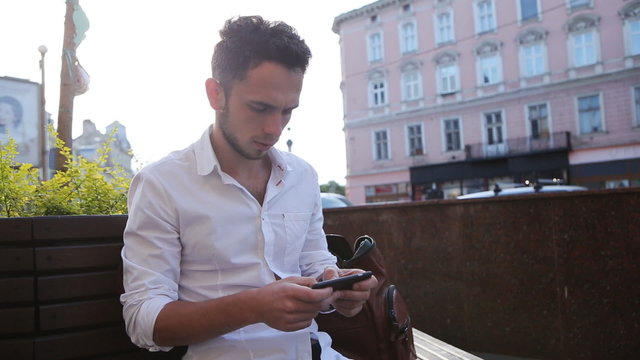Attractive young man sitting in a square and using his mobile phone for texting. Male in his 20s creating an sms on smartphone