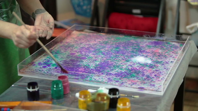 Red oil-based inks splashing over purple and green colors in a tank with water being prepared for marbling. The Ebru is a method of aqueous surface design
