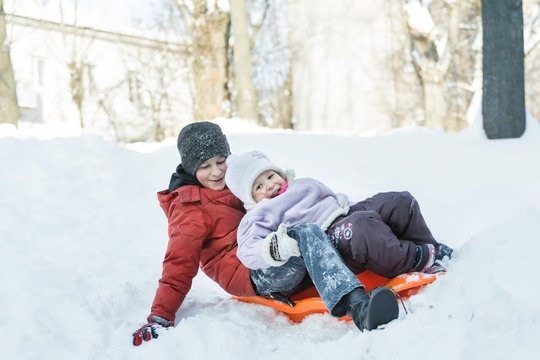 Sibling children laughing and riding downhill on winter orange toboggan sledge made of plastic outdoor