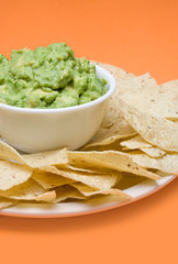 Guacamole and Chips – A bowl of fresh guacamole with corn tortilla chips. Bright orange background.