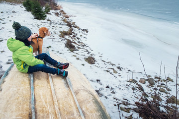 Boy with dog sitting together near frozen lake