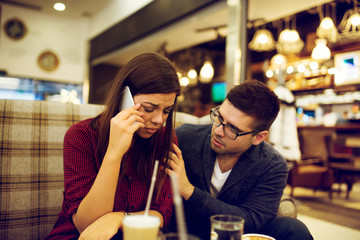 Couple at cafe, she is talking on mobile phone hearing bad news while he is consoling her.