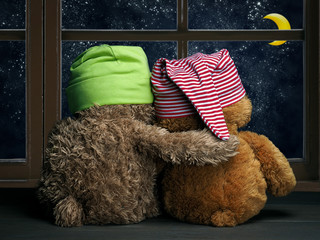 Two friends or lovers look out the window at night, the stars and the moon. Toys colorful hats cubs I sit embracing on a window sill and look out the window. Concept - love, friendship, support