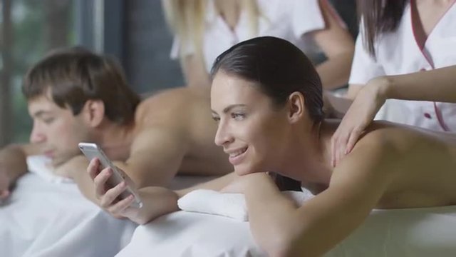 Young woman is using a smartphone during  a relaxing couple massage from female masseurs in wellness center. Shot on RED Cinema Camera.