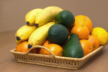 Still life with tropical fruits: bananas, avocados, lemons, oranges and tangerines lie on a brown wicker tray. Photo closeup