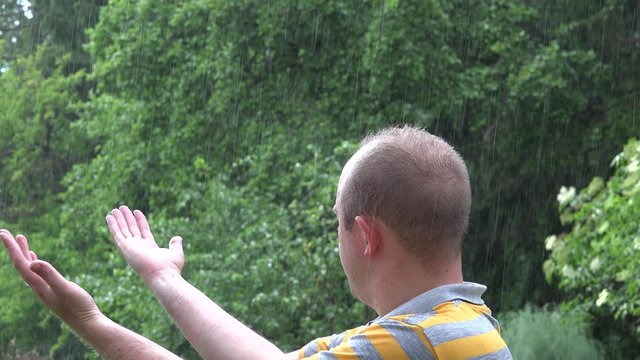 young happy thinking man enjoy summer rain in green park and raise hand up. 4K UHD video clip.  
