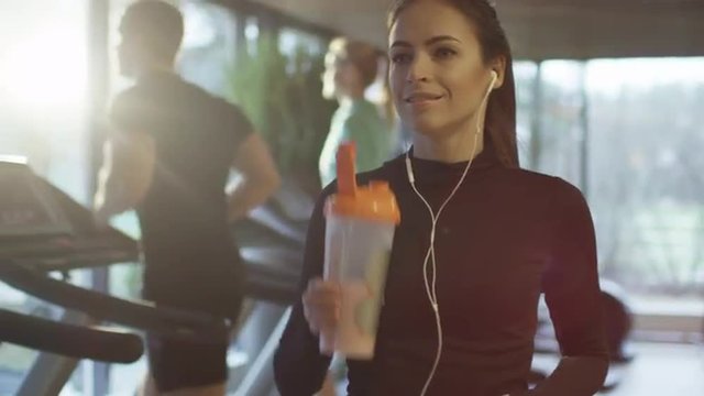 Attractive caucasian girl is drinking a protein shake drink next to a treadmill in the sport gym. Shot on RED Cinema Camera.