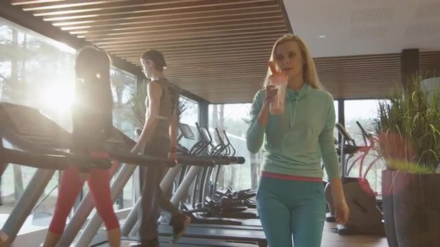 Attractive blond caucasian girl is drinking a protein shake drink while walking next to a treadmill in the sport gym. Shot on RED Cinema Camera.