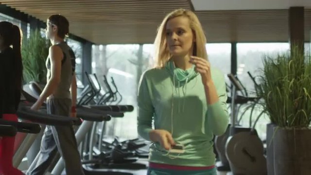 Attractive caucasian girl is starting to run on the treadmill in the sport gym with earphones. Shot on RED Cinema Camera.