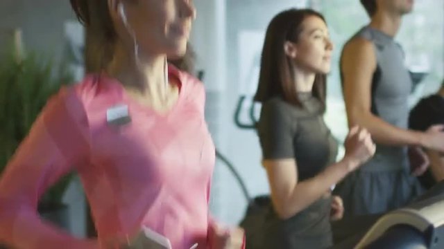 Slow motion footage of attractive caucasian girl running on the treadmill in the sport gym with earphones. Shot on RED Cinema Camera.