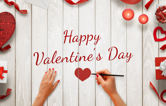 Girl hand painting Happy Valentines day message on a wooden background. Love decorations beside, gifts, candles, hearts