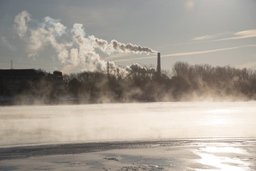 Steam rises off the water of the river and smoke and pollution puff out of a large coal smokestack on a cold winter day.