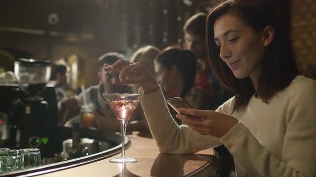 Attractive caucasian woman with a cocktail is using a smartphone in a bar. Shot on RED Cinema Camera.