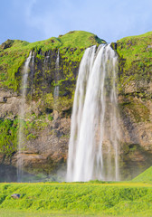 silver waterfall in sun shining day with green grass and blue sky in Iceland