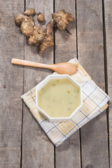 Top view of served artichoke soup in white bowl on plaid dishcloth with wooden spoon and raw artichokes on wooden background