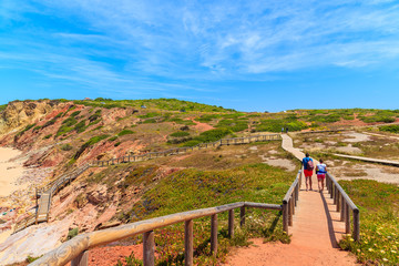 Couple of tourists walking on footbridge to Praia do Amado beach, famous place for surfing, Algarve region, Portugal
