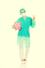 Female surgeon doctor with piggy bank shows victory sign