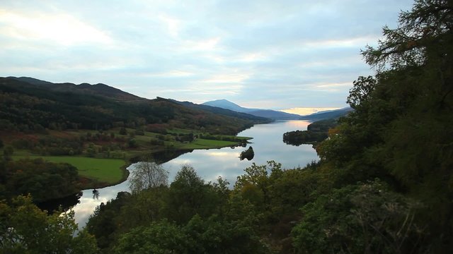The sun sets over a beautiful Scottish lake with rolling green hills in the background.