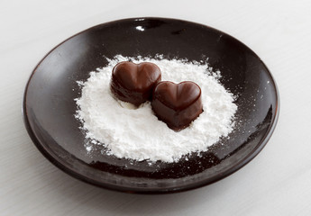 Two chocolate heart-shaped candies on a brown plate with sugar powder against wooden background
