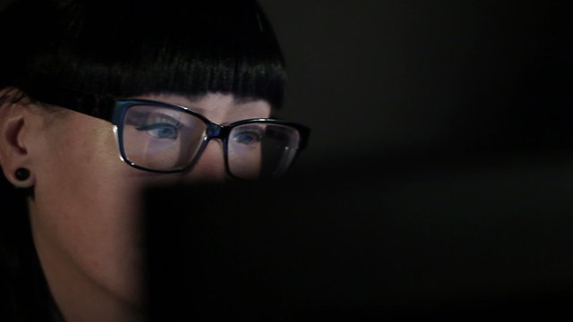 Girl in glasses sitting in front of the monitor and look at the screen. On linzae glare from the monitor. Black background. Close-up. Horizontal movement
