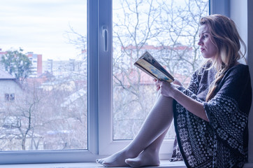 Young beautiful girl student sitting on a window sill at the window overlooking the city and thoughtfully reading a book a textbook with folk tales