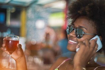 Young happy smiling black girl in sunglasses talking on the phone with a drink in the other hand, on a sunny day